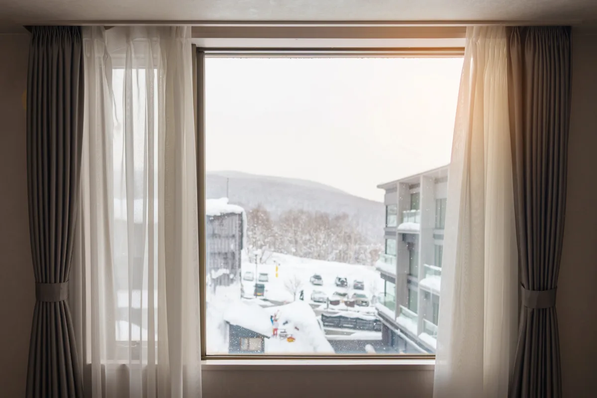 View through a window with sheer curtains, overlooking a snow-covered parking lot and apartment building, with snow-dusted hills in the background.