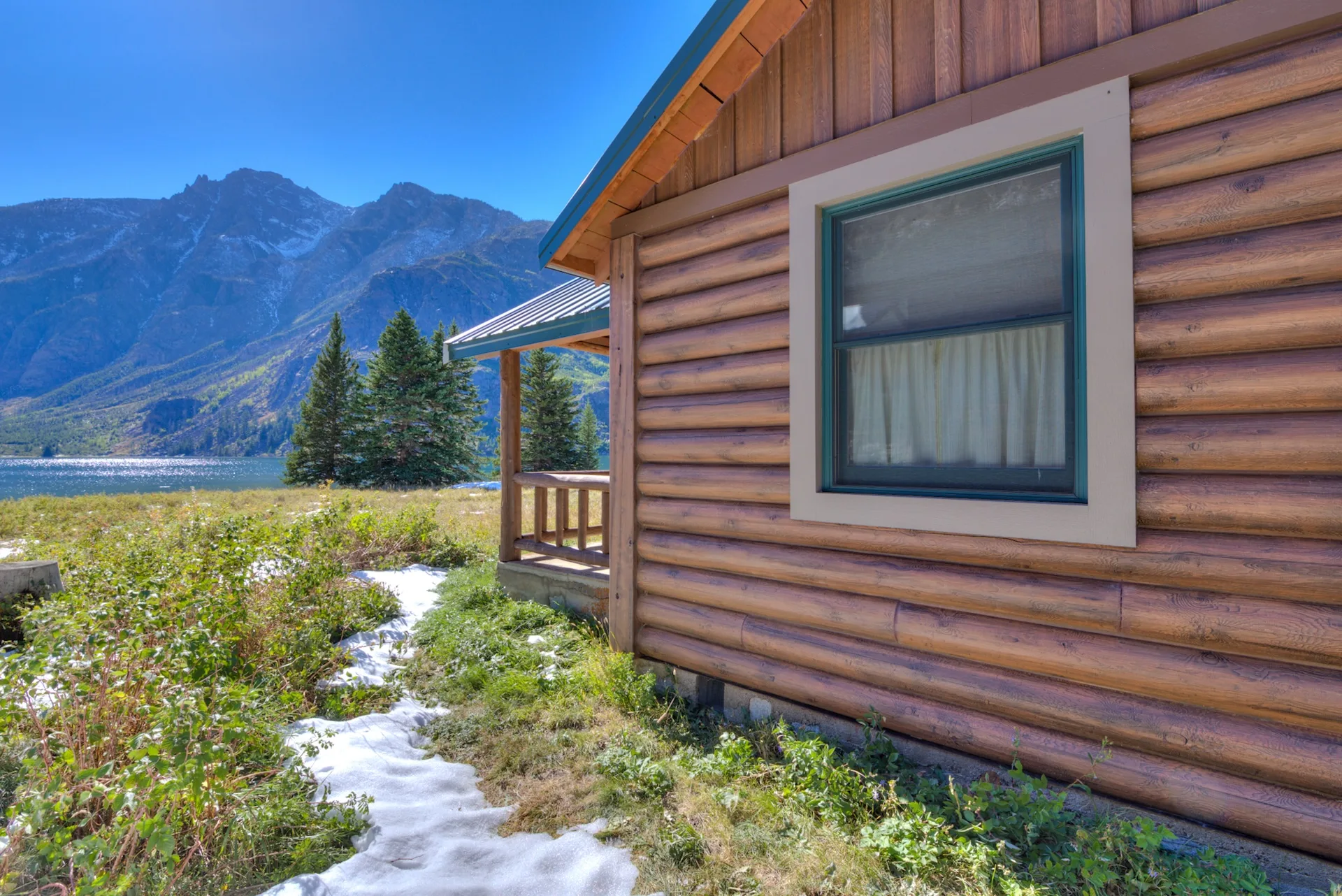 Rustic log-sided mountain home with a large window, surrounded by snow-covered ground, trees, and mountains in the background.