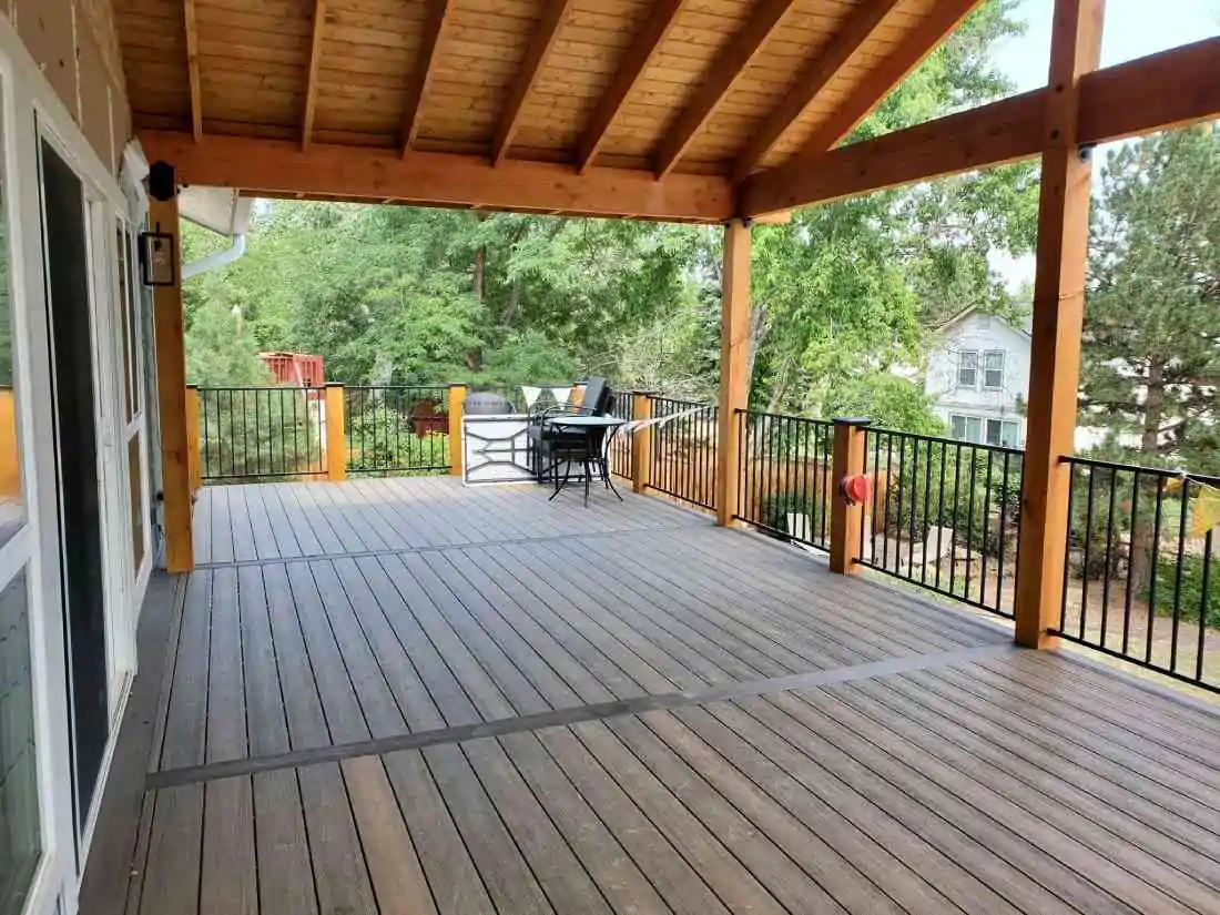 Covered wood deck with a black metal railing, seating area, and pergola-style roof, surrounded by trees with a house visible in the background.